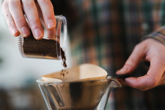 A person's hands pour freshly ground coffee from a small clear container into a paper filter set in a glass pour-over coffee maker. This is part of a morning routine.