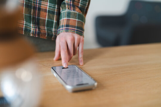 A person wearing a plaid shirt gently presses a finger onto the screen of a smartphone. The device rests on a light wooden surface, indicating active digital interaction.