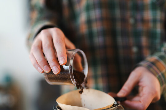 A person in a plaid shirt pours ground coffee from a small container into a paper filter for a pour-over coffee. Preparing a fresh morning brew.