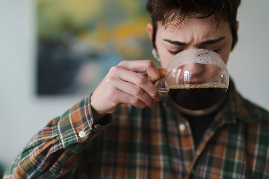 A young person in a plaid shirt sips dark coffee from a clear glass mug. Their eyes are closed, reacting to the strong, hot beverage. This captures a moment of morning intensity.