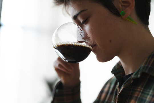 A young person with short hair and an ear taper drinks a dark beverage from a clear glass. Their eyes are closed, savoring the taste and aroma of the drink.