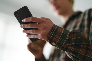 A person in a plaid shirt holds a black smartphone, actively engaging with the device. Their hands are visible, typing or swiping on the screen, focused on digital interaction.