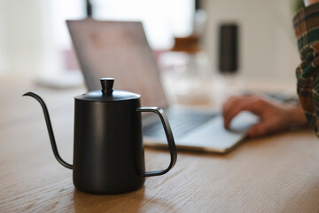 A person in a plaid shirt is focused on their laptop at a wooden desk. A sleek black gooseneck kettle sits nearby, suggesting a moment for a warm beverage.