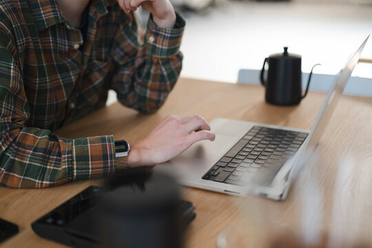 A person in a plaid shirt works on a laptop at a wooden desk. Their hand is on the trackpad, focused on the screen. A black coffee pot sits nearby.