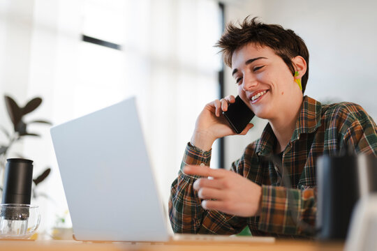 A happy young person with short hair is talking on a smartphone and pointing at a laptop screen. They are working remotely from a bright home office, multitasking efficiently.