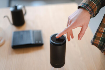 A person's hand presses the power button on a sleek black coffee grinder. A gooseneck kettle and digital scale are on the wooden table, ready for brewing.