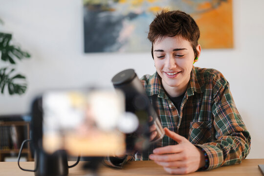 A smiling young person with short hair and earbuds records a video with a smartphone. They hold and present a black device, possibly a microphone or speaker.