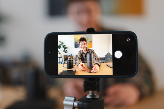 A smiling young person grinds coffee beans, being recorded by a smartphone on a tripod. The phone's screen shows the live camera view of the person at home.