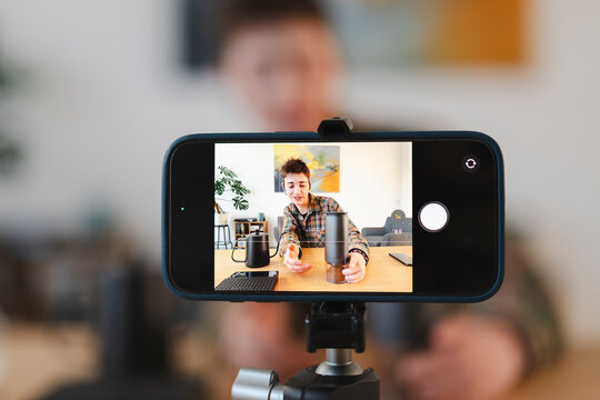 A young person records a video tutorial about coffee preparation. A smartphone on a tripod captures the live feed of the person holding a coffee grinder. This shows modern content creation.