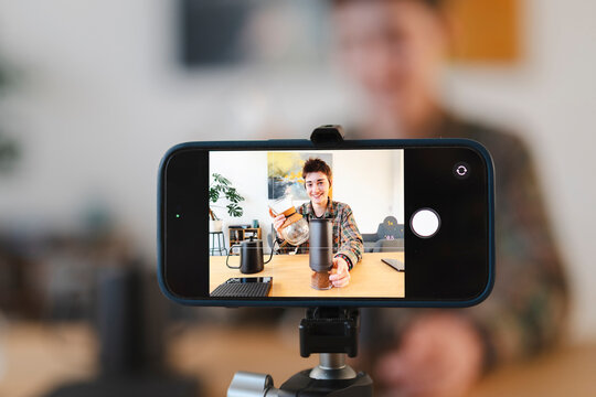 A young person records a coffee preparation tutorial or content using a smartphone on a tripod. They smile, holding a coffee grinder and carafe, showcasing the pour-over process.