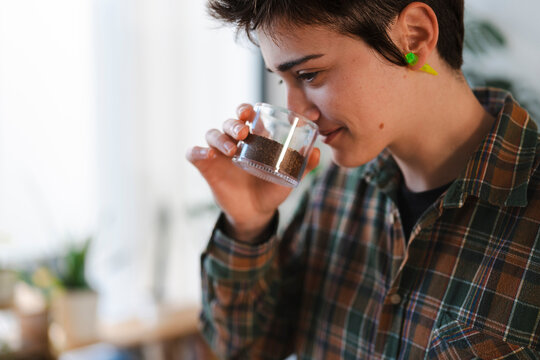A young person with short hair and a plaid shirt holds a small glass of ground coffee, smelling its aroma intently. They are focused on the sensory experience.