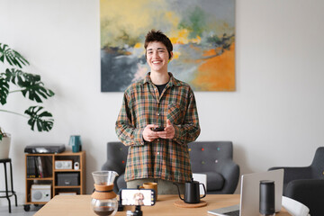 A young person smiles, holding a phone in a modern home office. Coffee brewing equipment, a laptop, and a recording smartphone are on the table.