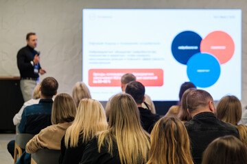 Fototapeta premium Speaker presents information to seated audience at corporate seminar. Large screen shows colorful diagrams. Attentive attendees listen, learn, engage with presenter at professional event.