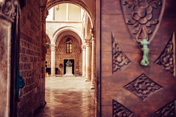 View of elaborately carved wooden door leads to a sun-drenched courtyard, revealing a statue framed by arched columns, Dubrovnik, Dubrovnik-Neretva County, Croatia.