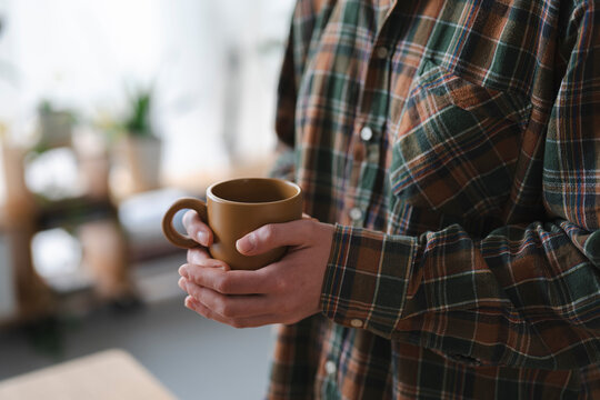A person wearing a comfortable plaid shirt holds a warm brown ceramic mug with both hands. The blurred background suggests a relaxed indoor setting, perfect for a quiet moment.