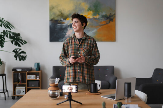 A smiling young person in a plaid shirt holds a phone while standing in a bright room. A smartphone on a tripod records a coffee setup on the table. They are creating online content.