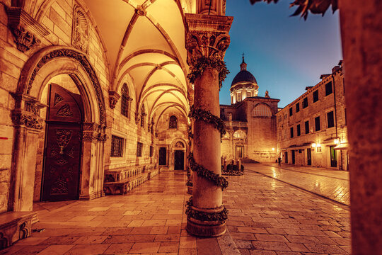 View of warmly lit stone arches and columns adorned with festive garlands frame a historic square under a twilight sky, Dubrovnik, Dubrovnik-Neretva County, Croatia.