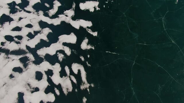 Top-down aerial shot of a frozen lake surface with dark cracked ice contrasting with white snow patches creating winter texture