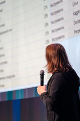 Presenter holds a microphone facing a large screen filled with charts and numbers. A professional scene of a talk or seminar about data and business topics.