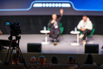 Panel discussion on stage with camera recording audience during conference event. Professional atmosphere as speakers share insights before attentive viewers.