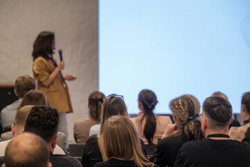 Presenter speaks to seated audience in bright conference room during training session. Attentive listeners take notes as slides fill large screen.