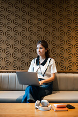 A young woman sits on a sofa in a trendy cafe, using a laptop, with a geometric patterned wall in the background.