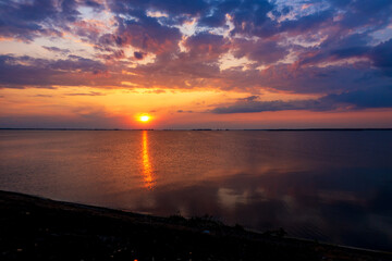 Fototapeta premium Vibrant sunset over a calm lake with golden sun reflection. Dramatic clouds in fiery orange, purple, and blue hues create a picturesque, serene natural landscape.