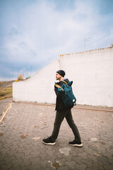 A stylish man with a teal backpack walks along a paved path next to a white wall under a cloudy sky, exuding a sense of urban adventure and casual exploration.