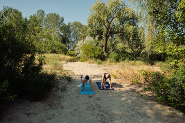 Fototapeta premium Two people practice yoga outdoors on sandy ground amidst lush green trees under a clear blue sky. This serene scene captures a healthy lifestyle and connection with nature.