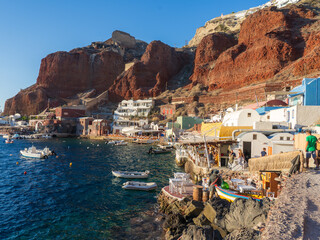 View of stark, red cliffs meet the turquoise sea, framing whitewashed buildings and boats dotting the harbor under a crisp blue sky, Oia, Thira, Greece.