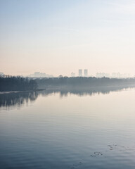 Serene morning river landscape with calm water reflecting a hazy sky and dark trees on the island. Silhouetted skyline of a city visible through the mist.