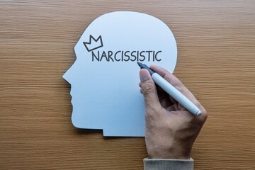 A hand writes NARCISSISTIC with a crown inside a paper head profile. Ego, self-importance, personality disorders concept.