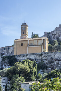 View of a sun-drenched church with a clock tower stands proudly atop a rocky hill, framed by lush greenery and the azure sky, Eze, Provence-Alpes-Cote d'Azur, France.
