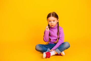 Naklejka premium Portrait of a young girl with glasses sitting cross legged on a bright yellow background wearing a purple turtleneck and jeans showing playful childhood style and curiosity