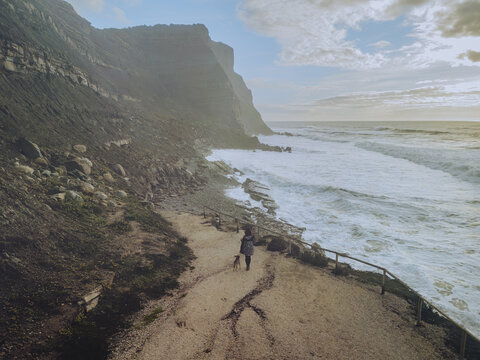 Aerial view of waves crashing against the rugged coastline near Praia do Porto Barril, with a lone figure walking along the path, Assenta, Lisbon District, Portugal.