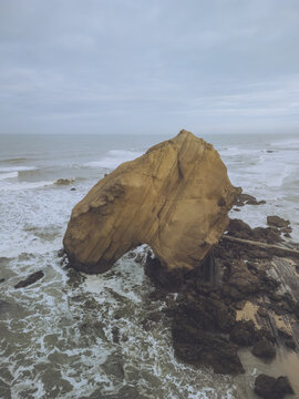 Aerial view of the monolithic rock formation standing defiantly against the relentless ocean waves, its warm tones contrasting with the cool, gray sea, Santa Cruz, Lisboa, Portugal.