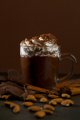 Hot chocolate with whipped cream and nuts served in a clear glass cup on a dark wooden table with chocolate and spices