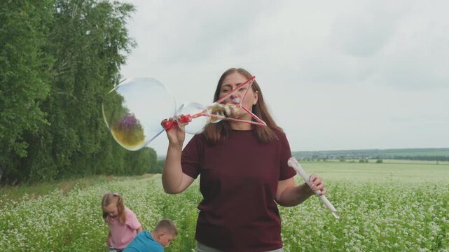 caucasian woman blowing bubbles in meadow with children playing behind her, birch tree line and cloudy sky framing casual family moment burgundy shirt, wand and iridescent soap orbs create playful