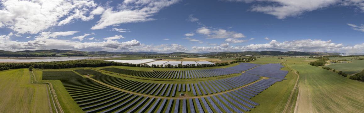 Aerial view of a vast solar farm glinting under a bright sky, contrasting with the lush green fields near the River Tay, Perth, Scotland, United Kingdom.