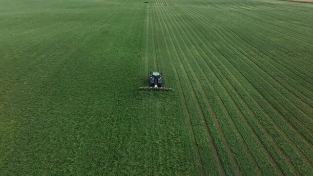 Top down view shows tractor moving forward on farmland, multi arm rake forming neat windrows on right while left remains uncut, cool tones and orderly striping