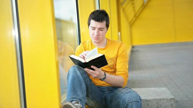 Young Man Sitting Outdoors Reading Holy Bible against Yellow Wall Background