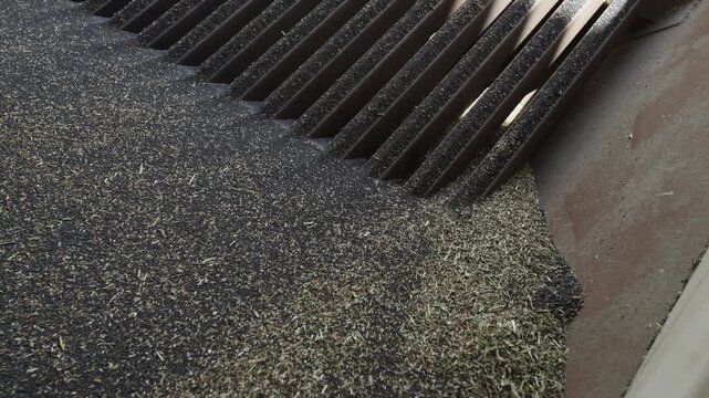 Slotted metal grate receives fine dark granules and grass clippings next to rusted steel panel. Material drifts toward slots. Low angle, shallow depth of field.