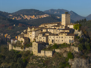 Aerial view of ancient stone buildings cluster atop a hill, crowned by a square tower against a backdrop of rolling mountains, Arrone, Umbria, Italy.