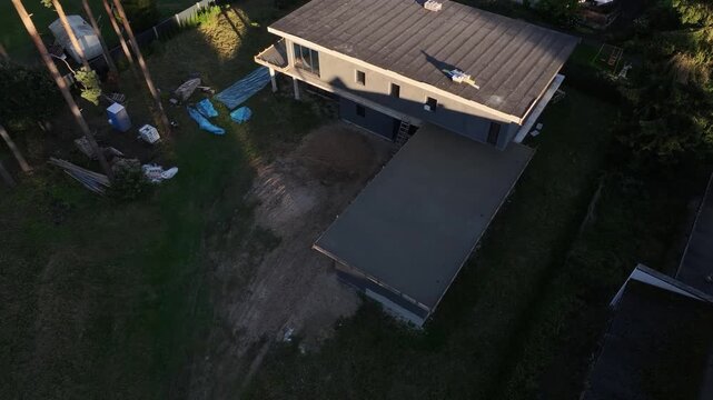 Aerial drone flyover of unfinished flat roof house and pine trees. Title in description. Late afternoon light, slow motion reveals roof details, tarps, slabs, and materials.