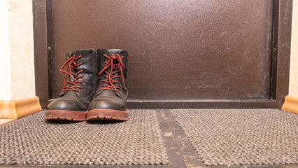 Worn lace up boots standing on dirty entrance mat in hallway, illustrating muddy footwear after bad weather, home cleaning routine