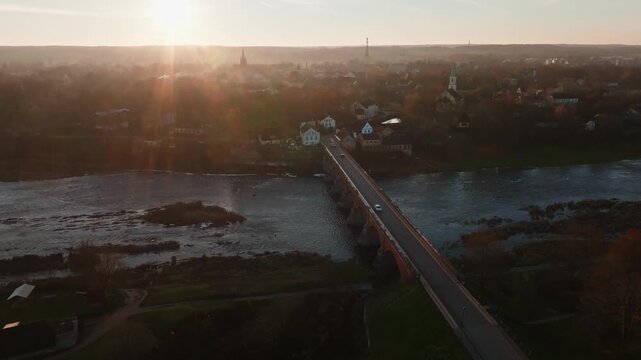 Drone view shows a narrow bridge over a shallow river toward a small European town. Vehicles move slowly. Lens flare, mist, and long shadows suggest autumn.