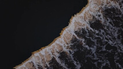 Aerial top down view shows waves breaking along unknown coast. Ochre sand ridges meet near black water. Foam threads shift as breakers move diagonally across frame. - Powered by Adobe