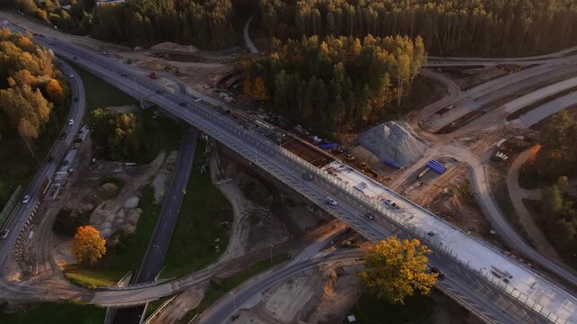 Drone follows diagonal bridge axis as traffic moves and crews work on new concrete decking, cloverleaf ramps, and trailers near suburban forest and gravel piles.
