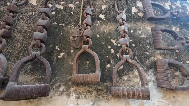 Collection of antique iron shackles, chains, and rusted hand tools hanging on a weathered stone wall in a heritage site