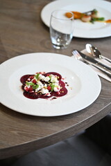 Close-up of a modern restaurant dish served on a white plate, featuring beet carpaccio topped with fresh greens, cheese, and garnish. Elegant food presentation on a wooden table with soft natural ligh